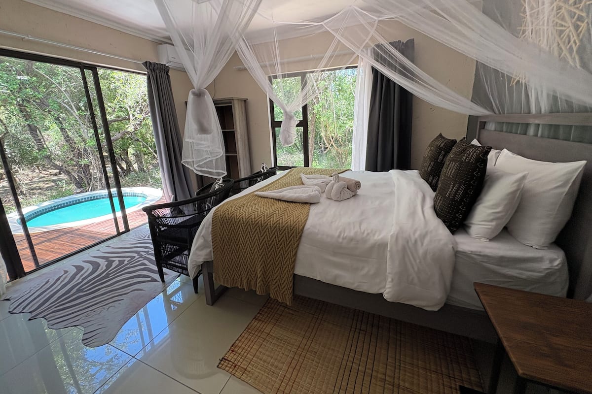 A bedroom with a bed, a bedside table, a window with a view of a swimming pool, and a mosquito net.