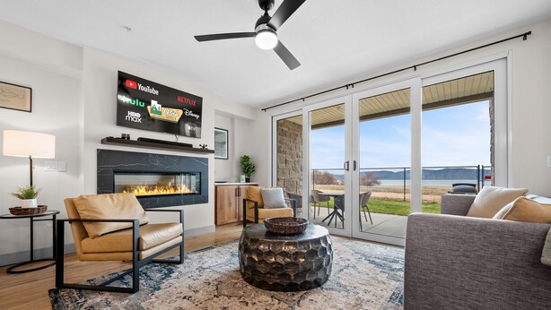 A living room with a fireplace, a flat-screen TV mounted on the wall, a ceiling fan, and a large sliding glass door leading to an outdoor patio. - Water's Edge 102 | Bear Lake Views| Lakeside Unit (Garden City)