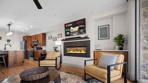 A modern living room with a fireplace, a kitchen area, and a mounted TV displaying streaming services. - Water's Edge 102 | Bear Lake Views| Lakeside Unit (Garden City)