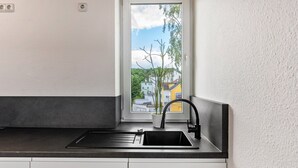 Uma cozinha com pia preta, armários brancos, janela com vista para edifícios e árvores, e um vaso com planta no parapeito.