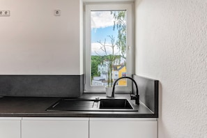 Una cocina con un lavamanos negro, gabinetes blancos, una ventana con vistas a edificios y árboles, y una planta en maceta en el alféizar.