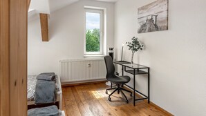 Quarto com piso de madeira, uma cama com cobertor cinza, um radiador, uma escrivaninha com cadeira e uma janela com vista para árvores.