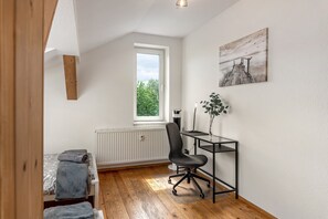 Un dormitorio con suelo de madera, una cama con manta gris, un radiador, un escritorio con silla y una ventana con vistas a árboles.