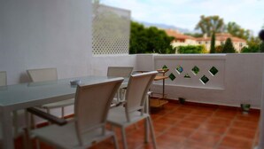 Um terraço com mesa e cadeiras, uma parede branca com painéis de vidro decorativos e vista para edifícios e árvores.