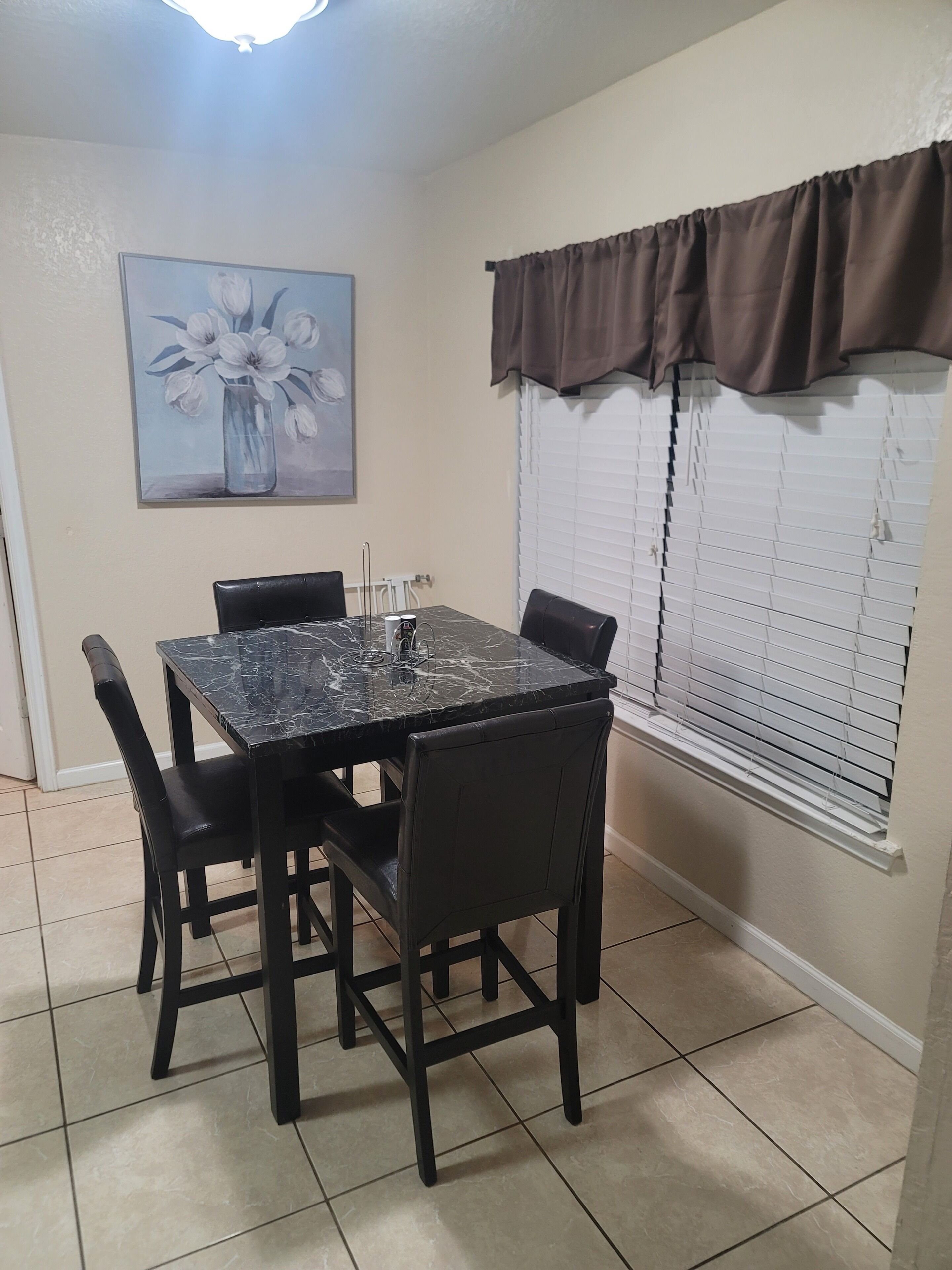 A dining area with a marble table, black chairs, a painting of flowers, and a window with blinds.