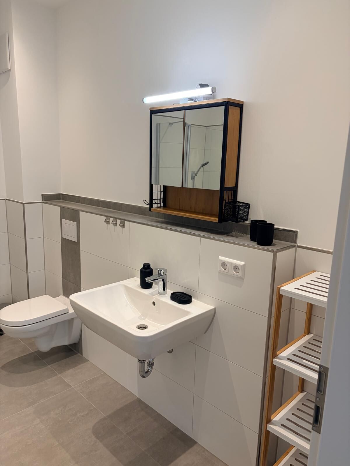 Un bagno moderno con un lavandino bianco, un water, uno specchio e uno scaffale con articoli da toilette.