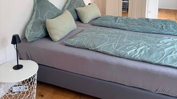 A modern bedroom with a grey bed, matching pillows, a white bedside table, and a wooden floor.