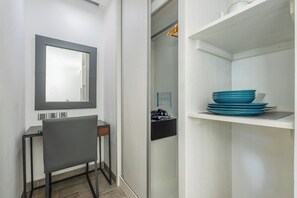 Une petite pièce comprenant un bureau, une chaise et un miroir. On y trouve une étagère avec des assiettes bleues et un petit objet noir posé dessus.