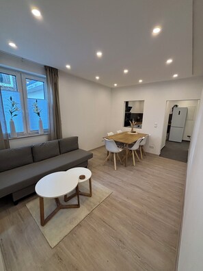 Ein modernes Wohnzimmer mit einer Couch, einem Esstisch mit Stühlen und einer durch eine offene Tür sichtbaren Küchenzeile.