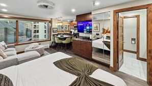 Una habitación de hotel moderna con una cama grande, un rincón de descanso con sofá y sillones, una kitchenette y un baño con espejo y puerta de madera.