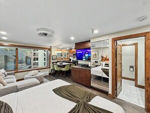 A modern hotel room with a large bed, a sitting area with a sofa and chairs, a kitchenette, and a bathroom with a mirror and wooden door. - Remodeled King Studio | AC | Ski-in/out | Peak 9 (Breckenridge)