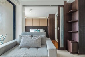 Ein modernes Schlafzimmer mit grauem Sofa, einem Bett mit weißen und türkisfarbenen Kissen, Holzregalen und einer Schiebeglastür zum Badezimmer.