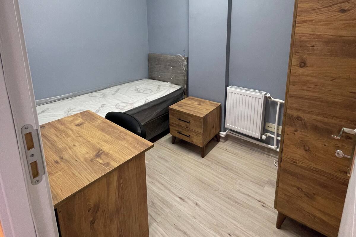 A compact room with a bed, wooden desk, and wardrobe, featuring a sloped ceiling and light-colored walls.