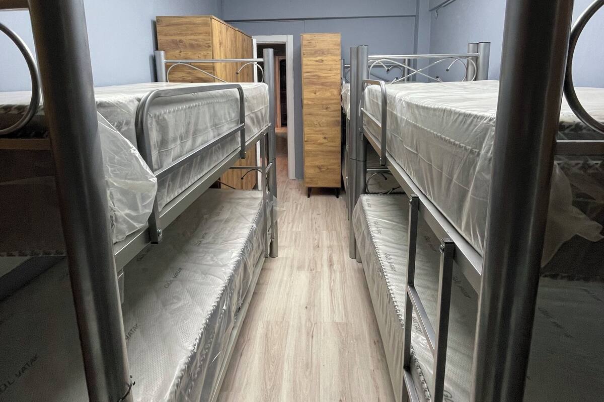 A dormitory room with bunk beds, a wooden wardrobe, and a door leading to another room.