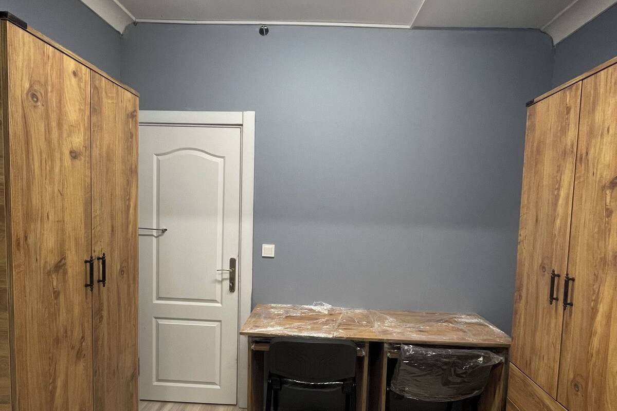 A room with a wooden wardrobe, a white door, a table with a plastic cover, and a chair.