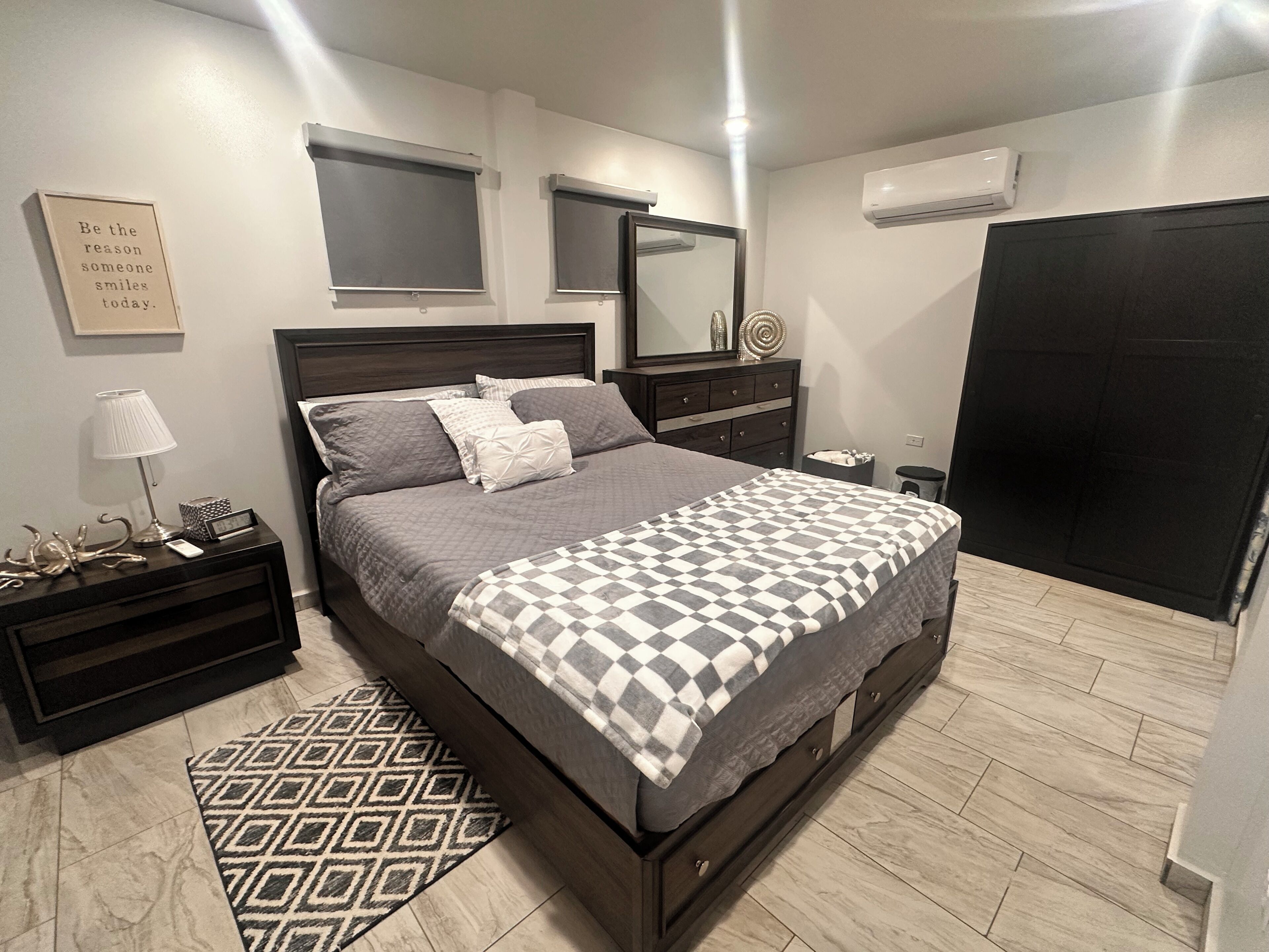 A bedroom with a bed, a nightstand, a lamp, a mirror, and an air conditioning unit.