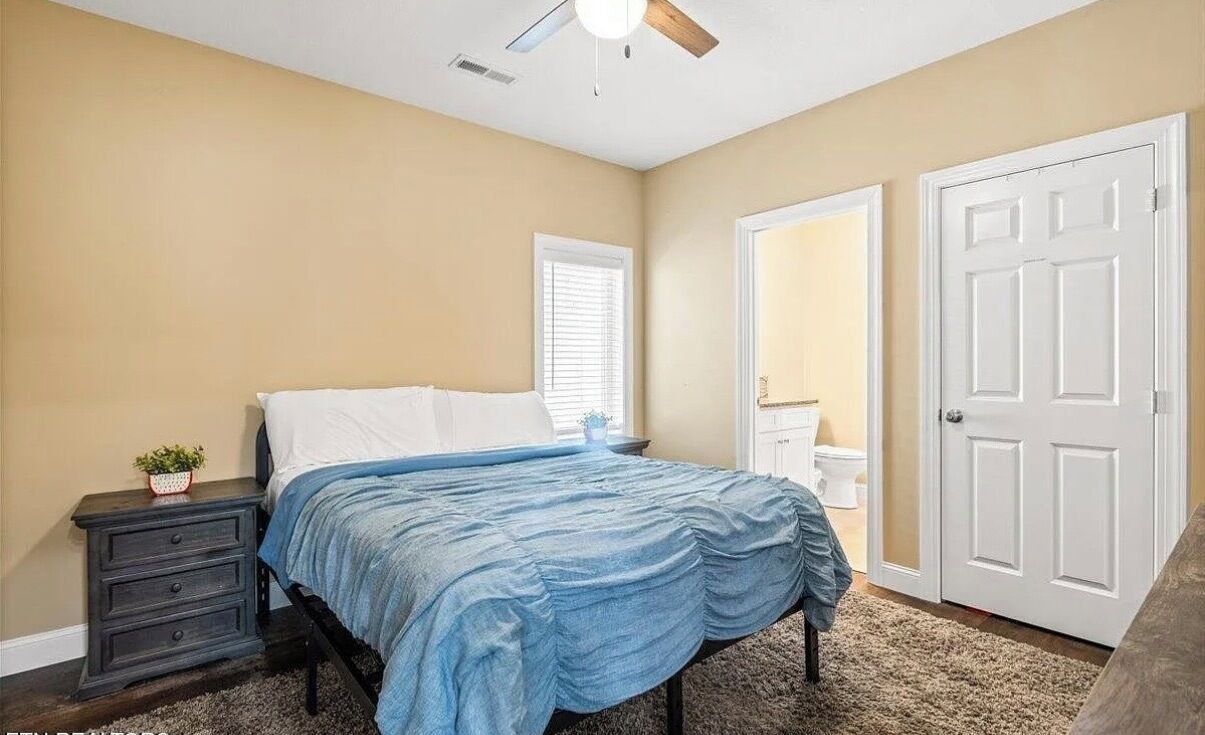 Ein Schlafzimmer mit einem Bett, einem Nachttisch, einem Deckenventilator, einem Fenster und einer Tür.