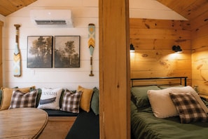 A cozy cabin interior with a wooden ceiling, a bed with green bedding, a round wooden table, and a bench with plaid cushions.