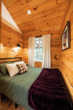 A wooden cabin bedroom with a bed, pillows, a blanket, a window with curtains, and a wall-mounted picture. - Henrys cabin on the river (Northbrook)
