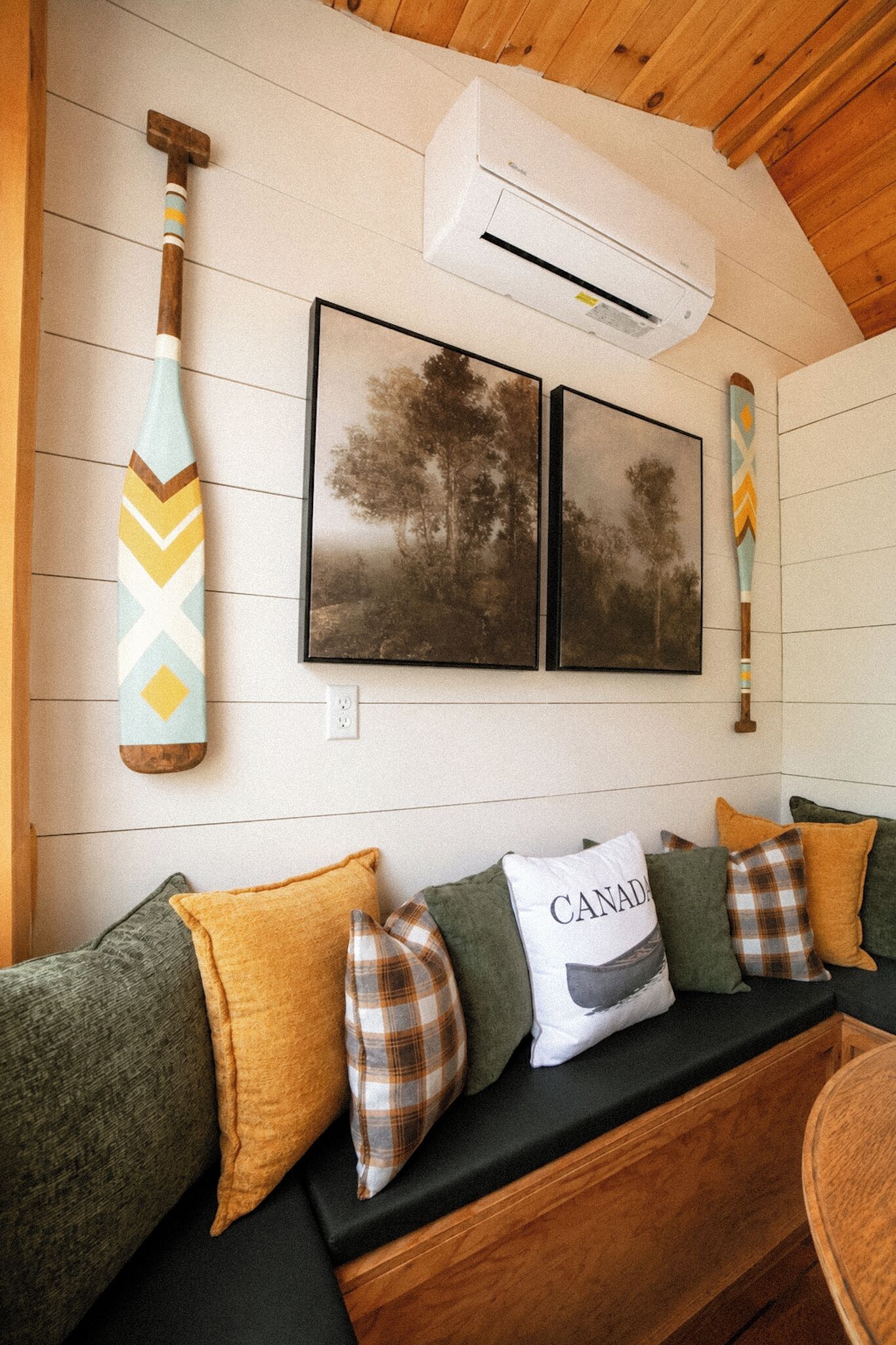 Una habitación con techo de madera, aire acondicionado instalado en la pared, dos cuadros enmarcados, un bate de madera y un sofá con varios cojines.