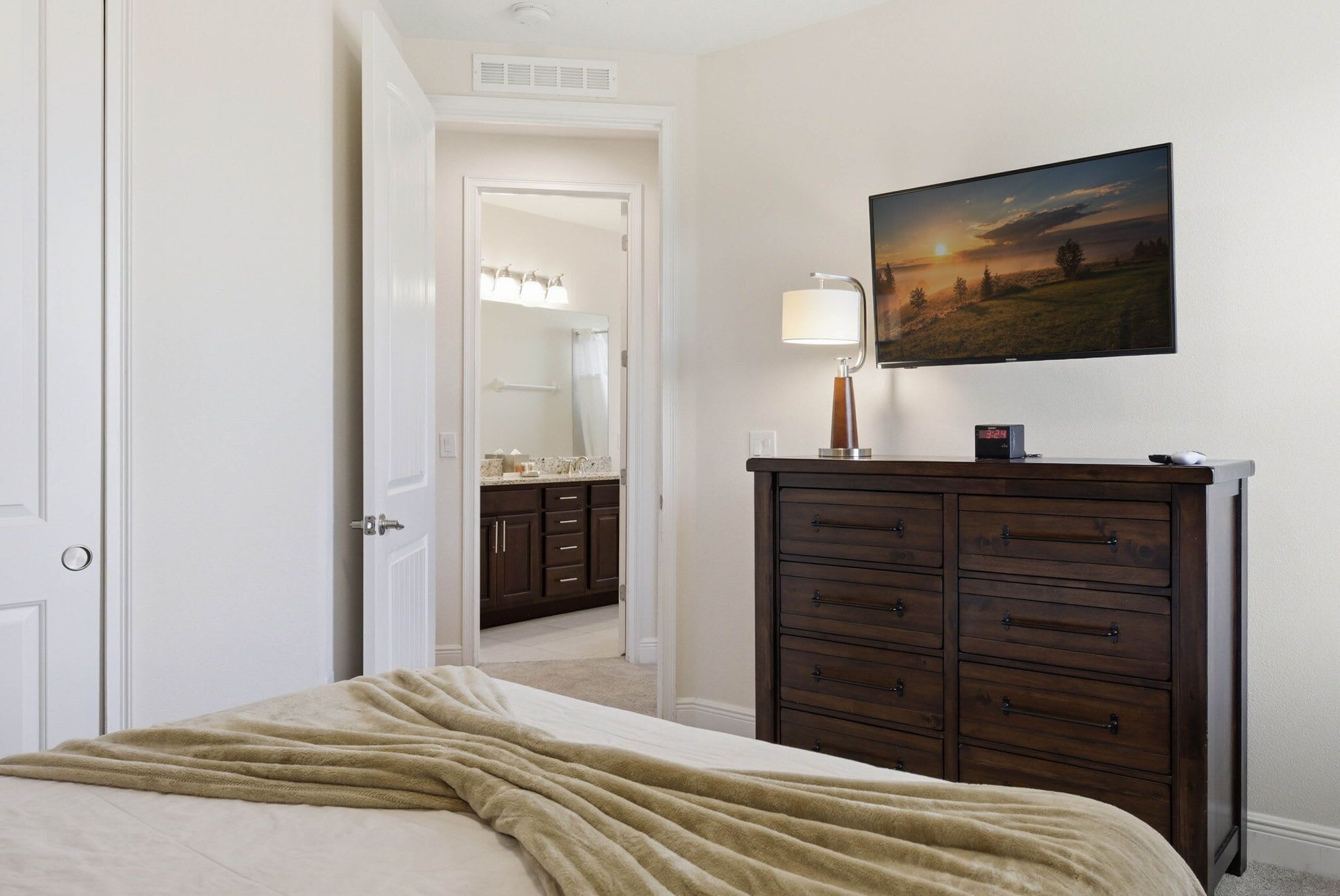 Un dormitorio con cama, cómoda, un cuadro y baño visible a través de una puerta abierta.