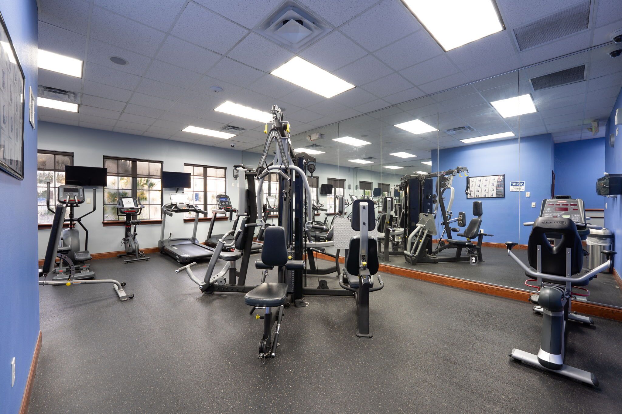 A gym with various exercise machines, a mirrored wall, and a window with blinds.