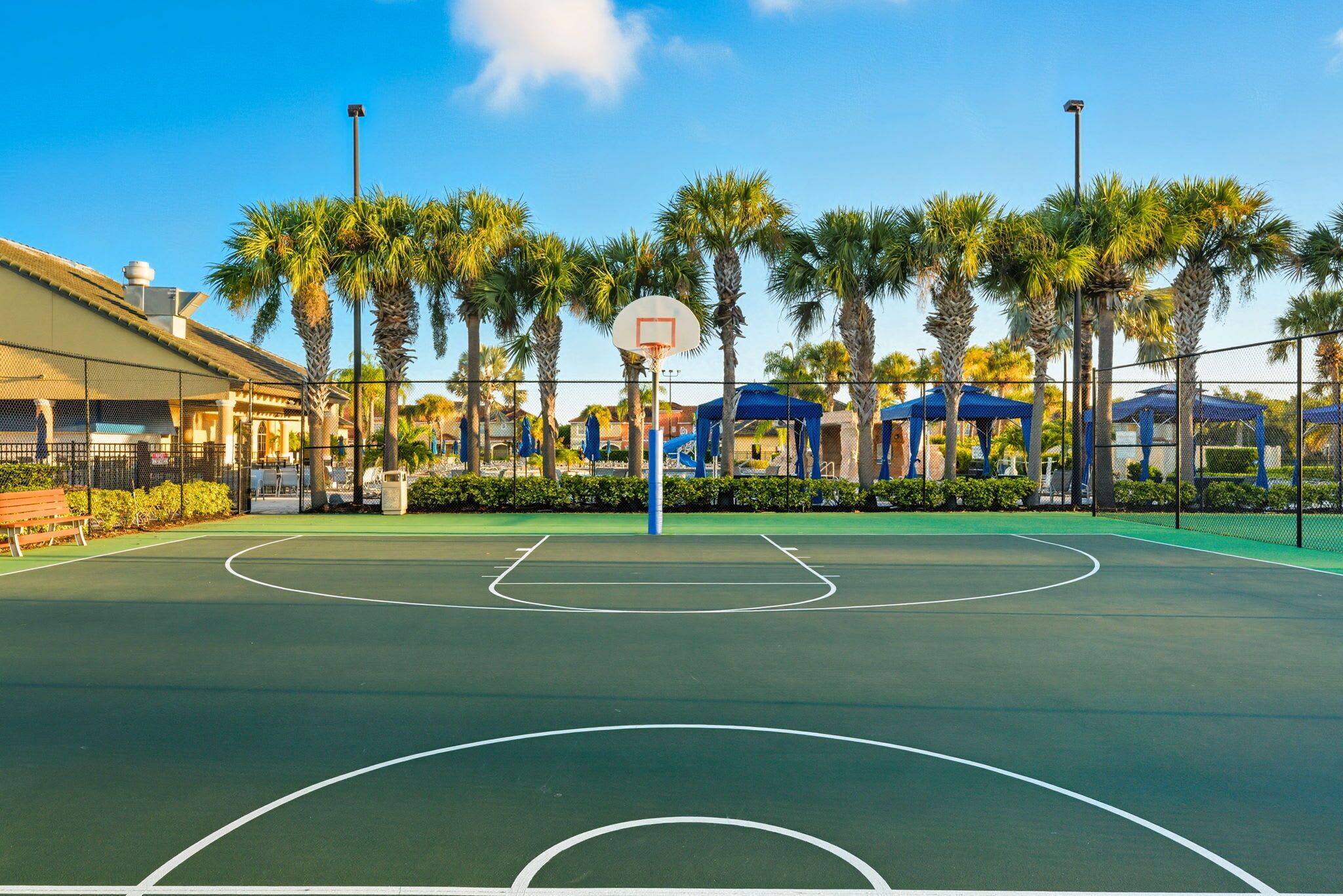 Una cancha de baloncesto al aire libre con aro, rodeada de palmeras y zonas de estar cubiertas.