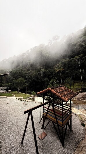 A playground with a swing set and a small shelter, surrounded by a gravel area and dense forest.