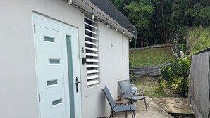 Uma casa com porta azul clara, um pequeno pátio com duas cadeiras, um vaso de planta e um caminho de concreto.