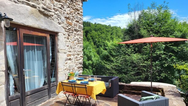 A stone building with a dining area featuring a table set for four, outdoor furniture, and a red umbrella. - Gîte de l'Escoutal 2/4 persons (LE BLEYMARD)