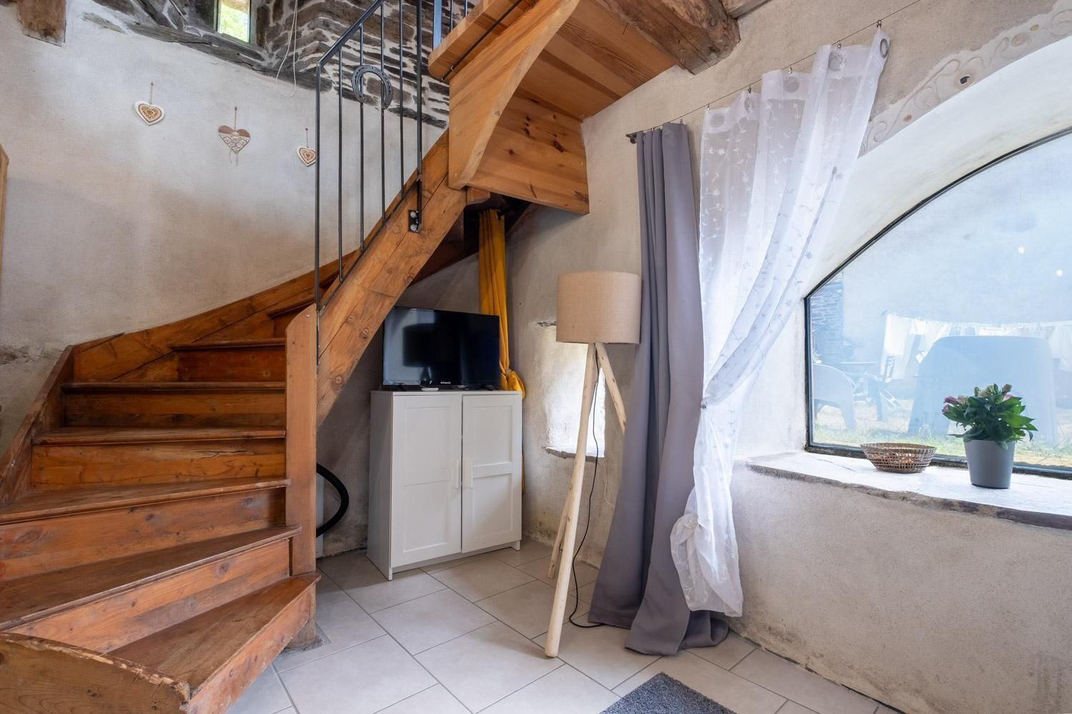 Una escalera de madera lleva a una pequeña cocina con televisión, lámpara y ventana con cortinas.