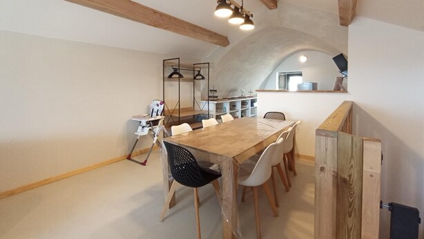 Un comedor con una mesa y sillas de madera, una lámpara colgante, y una zona de cocina con estantes y una ventana.