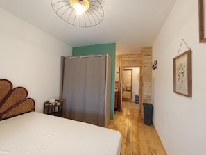 Quarto com cama, piso de madeira, ventilador de teto, porta que dá para outro cômodo e um quadro pendurado na parede.