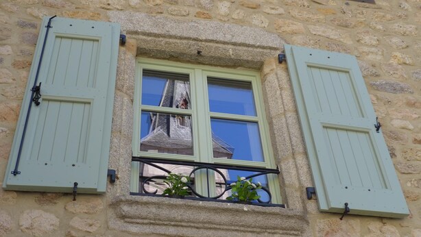 Un edificio de piedra con una ventana que tiene contraventanas verdes, un balcón con plantas y una torre visible al fondo.