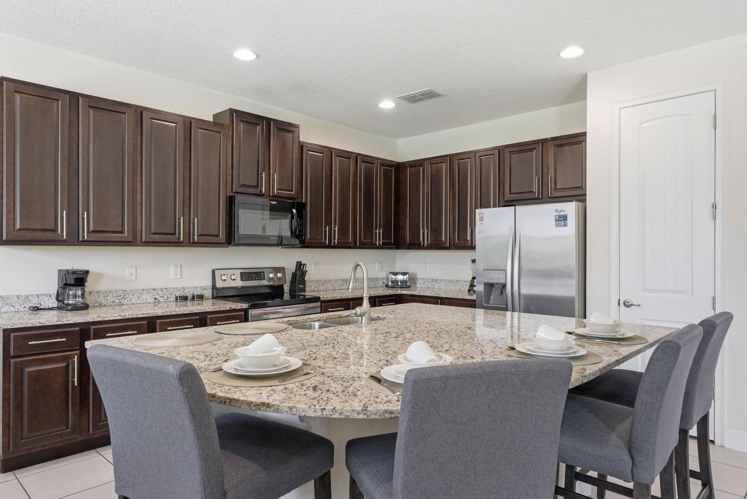 A modern kitchen with dark wood cabinets, granite countertops, a stainless steel refrigerator, and a dining area with four gray chairs.