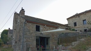 A stone house with a large window, a patio with a table and chairs, and a stone wall. - Gîte Lozère Bleue 4/5 persons (LES LAUBIES)