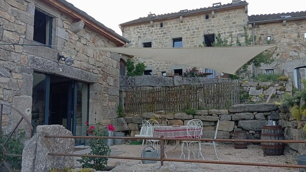 A stone building with a patio area featuring a table and chairs, a wooden fence, and a metal chair. - Gîte Lozère Bleue 4/5 persons (LES LAUBIES)