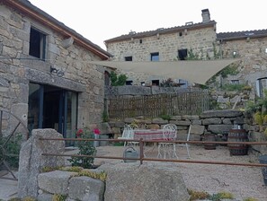 A stone building with a patio area featuring a table and chairs, a wooden fence, and a metal chair. - Lozère Bleue Vacation Rental for 4–5 People (LES LAUBIES)