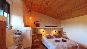A cozy bedroom with a wooden ceiling, a bed with a patterned bedspread, a chair, and a small table with a plant. - Gîte Lozère Bleue 4/5 persons (LES LAUBIES)