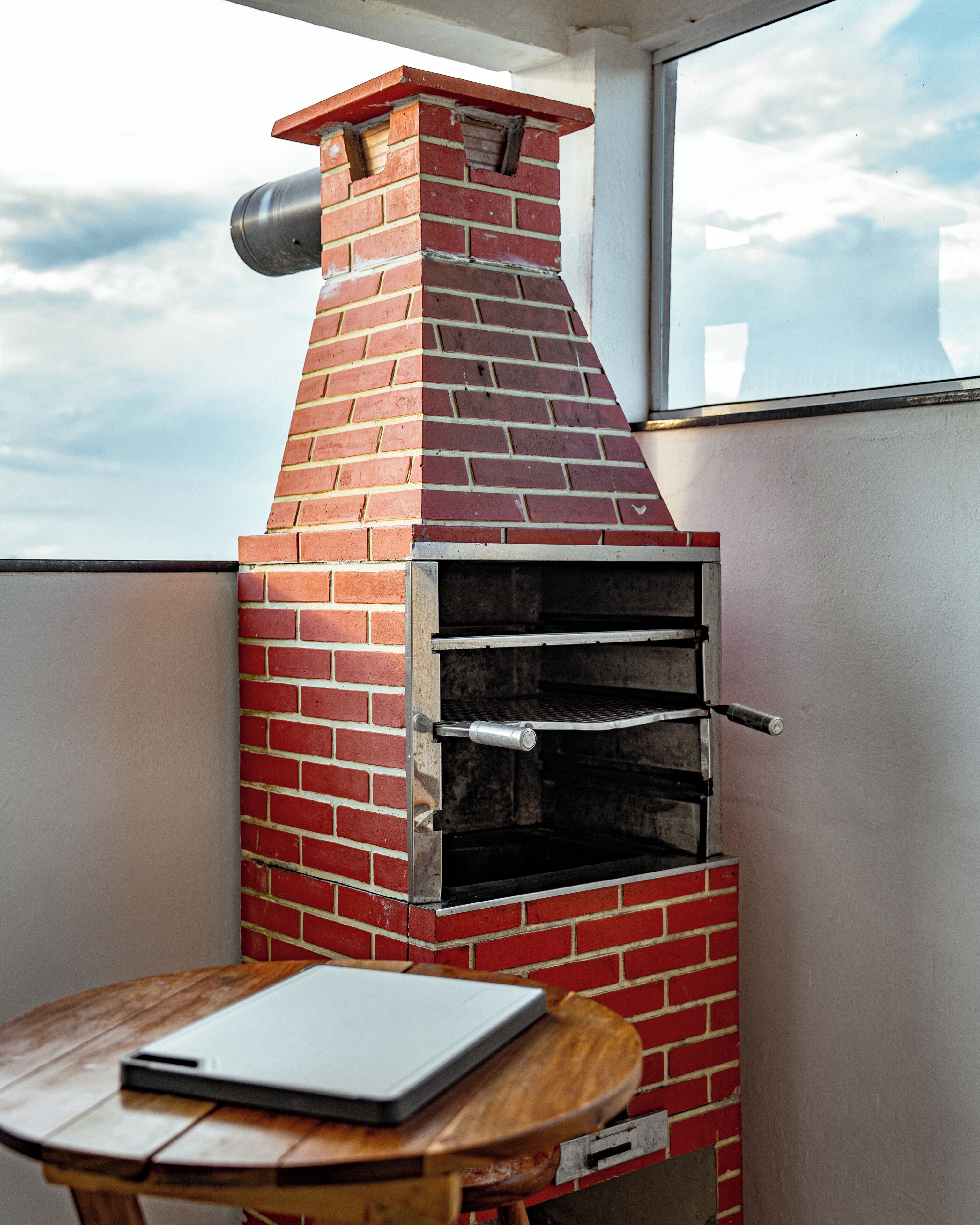 Un horno de ladrillo al aire libre con una chimenea metálica, una mesa con una computadora portátil y vistas al cielo a través de una ventana.