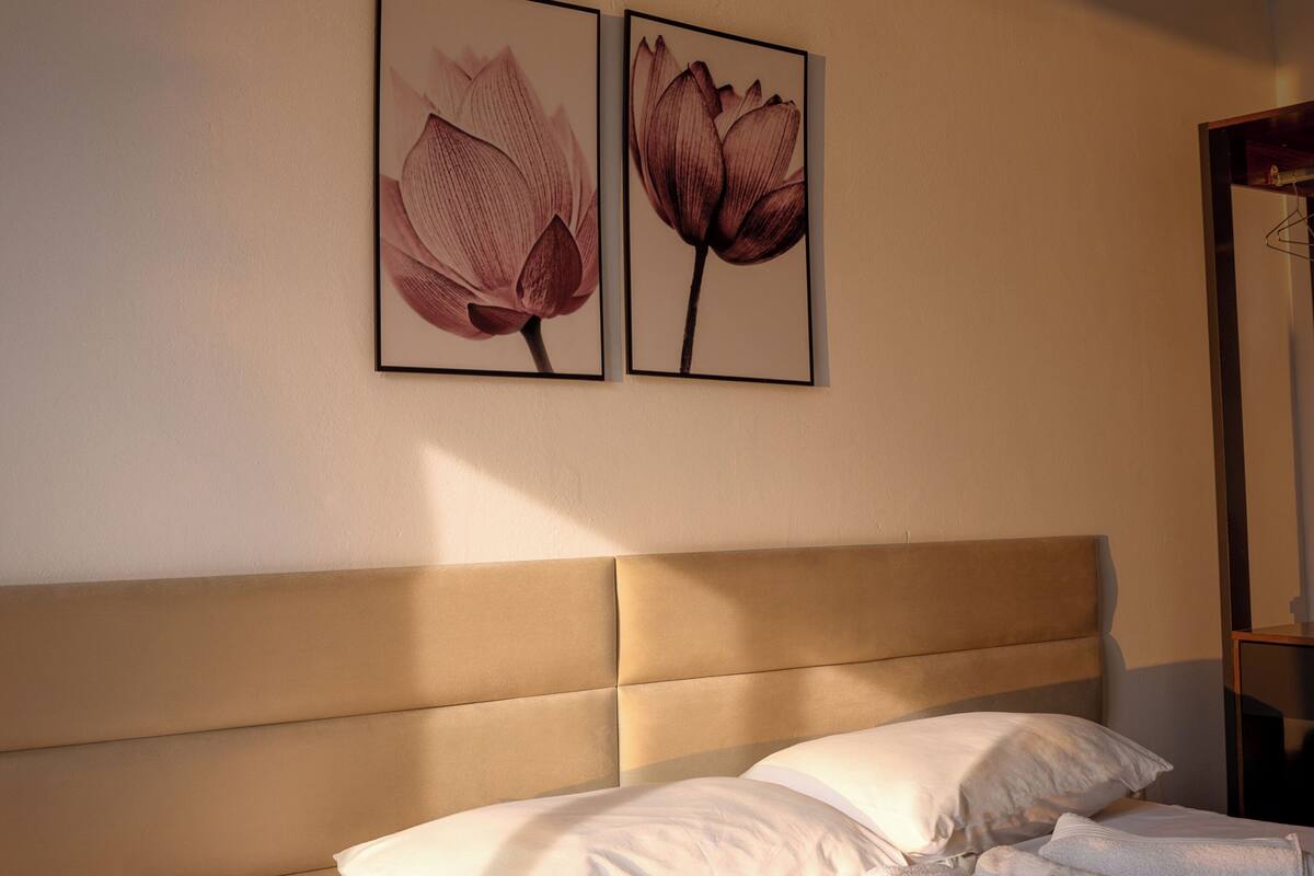Un lit bien fait, avec des oreillers et des serviettes, une tête de lit et deux tableaux représentant des motifs floraux, encadrés et accrochés au mur.
