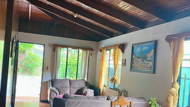 A spacious living area with a wooden ceiling, a grey sofa, a dining table with chairs, and a potted plant. - Marisol House (Quepos)