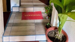 A wooden door, a red mat with 'BIENVENIDO' text, a potted plant, and a tiled floor. - Marisol House (Quepos)