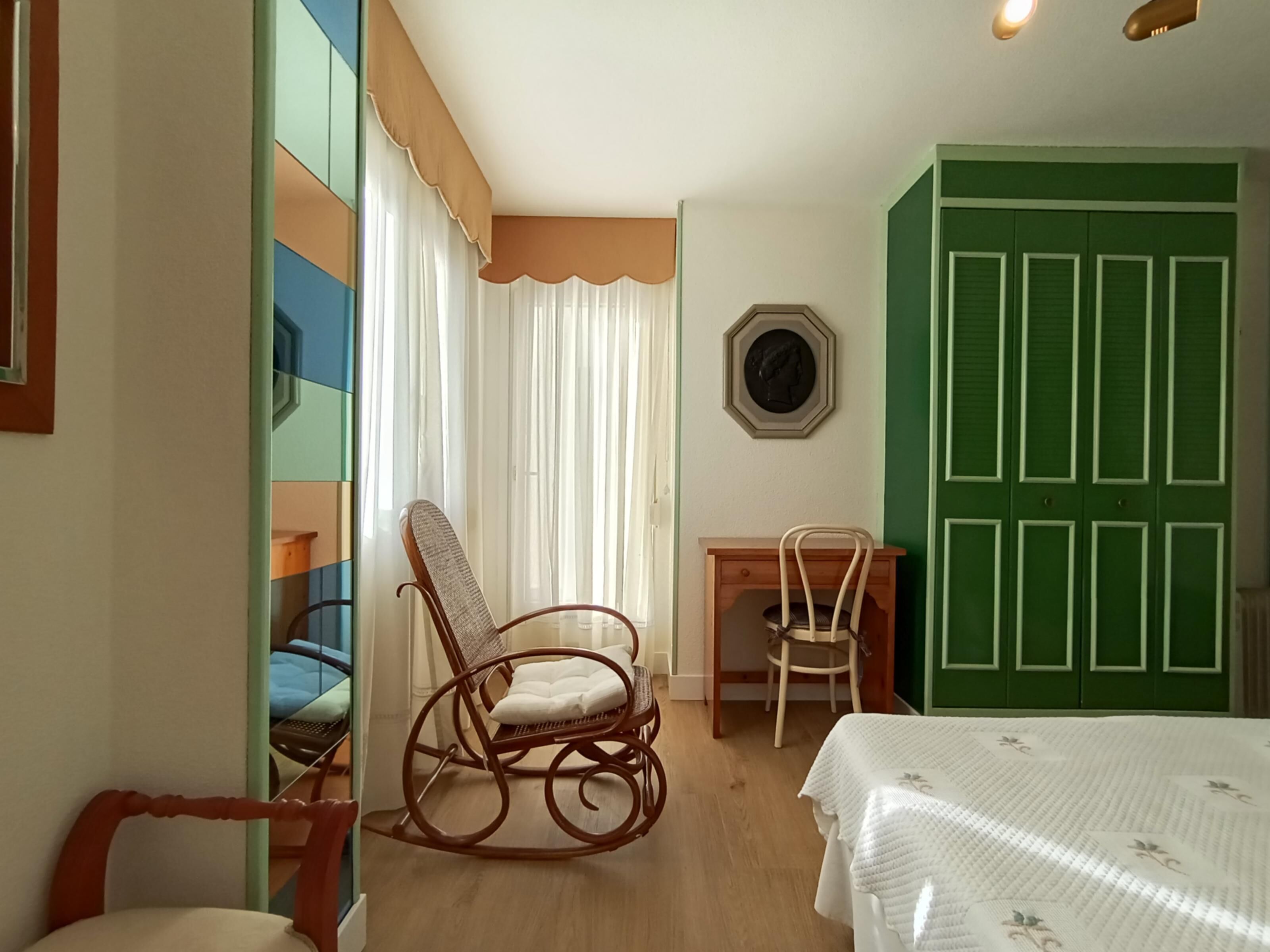 A room with a rocking chair, a small table, a green wardrobe, and a bed with a white bedspread.