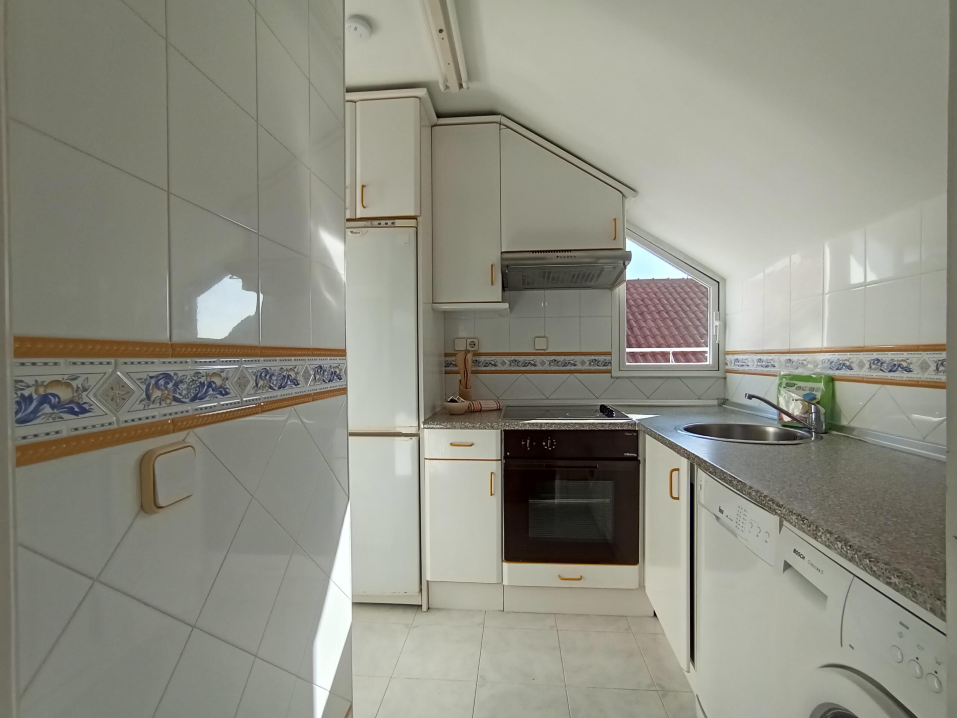 Cocina con armarios blancos, lavavajillas Bosch, horno y ventana con vista a tejados rojos.