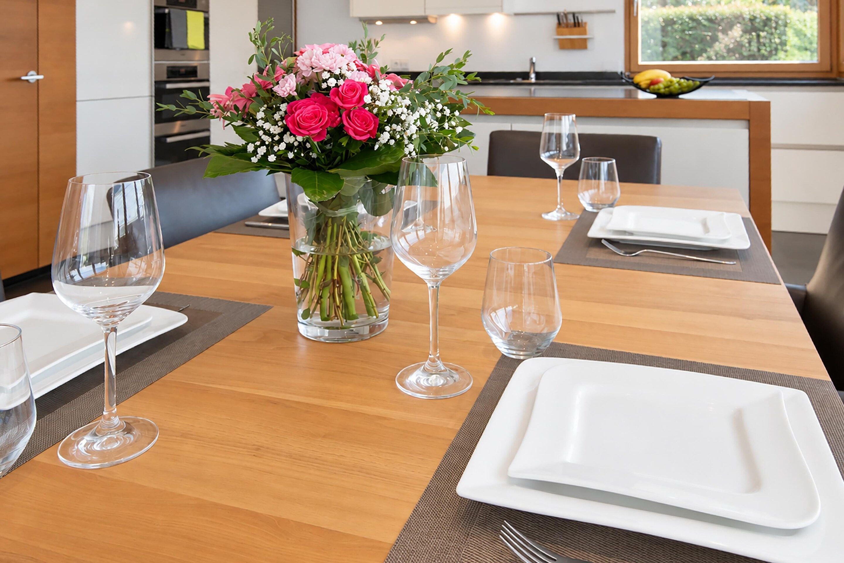 Uma mesa de jantar posta com copos de vidro transparente, pratos e um vaso com flores.