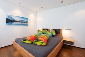 A modern bedroom with a wooden bed frame, a nightstand, a wall-mounted lamp, and a painting of a mountain landscape.