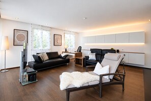 Una sala de estar moderna con un sofá de cuero negro, un otoman blanco y mullido, una silla de mimbre y un televisor de pantalla plana.