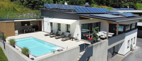 Ett modernt hus med en swimmingpool, solpaneler och en uteplats med solstolar.