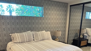 Quarto moderno com cama grande, papel de parede estampado, mesa de cabeceira com luminária e vista para árvores pela janela.
